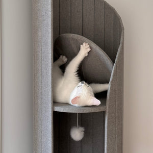 Cat lounging on padded platform inside Time Travel Tower Cat Tree in Light Grey, with dangling pompom toy.