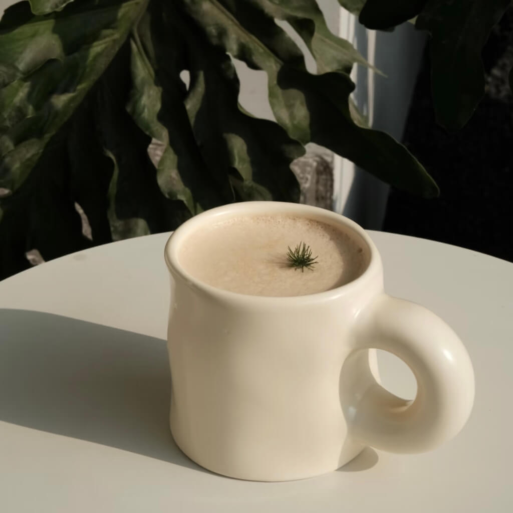 Pinch Ceramic Mug in Cream White, filled with a foamy latte topped with a minimalist green garnish, displayed outdoors with lush greenery in the background. A perfect match to the Meoof Elevated Pet Bowl for harmonious living.