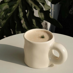 Pinch Ceramic Mug in Cream White, filled with a foamy latte topped with a minimalist green garnish, displayed outdoors with lush greenery in the background. A perfect match to the Meoof Elevated Pet Bowl for harmonious living.