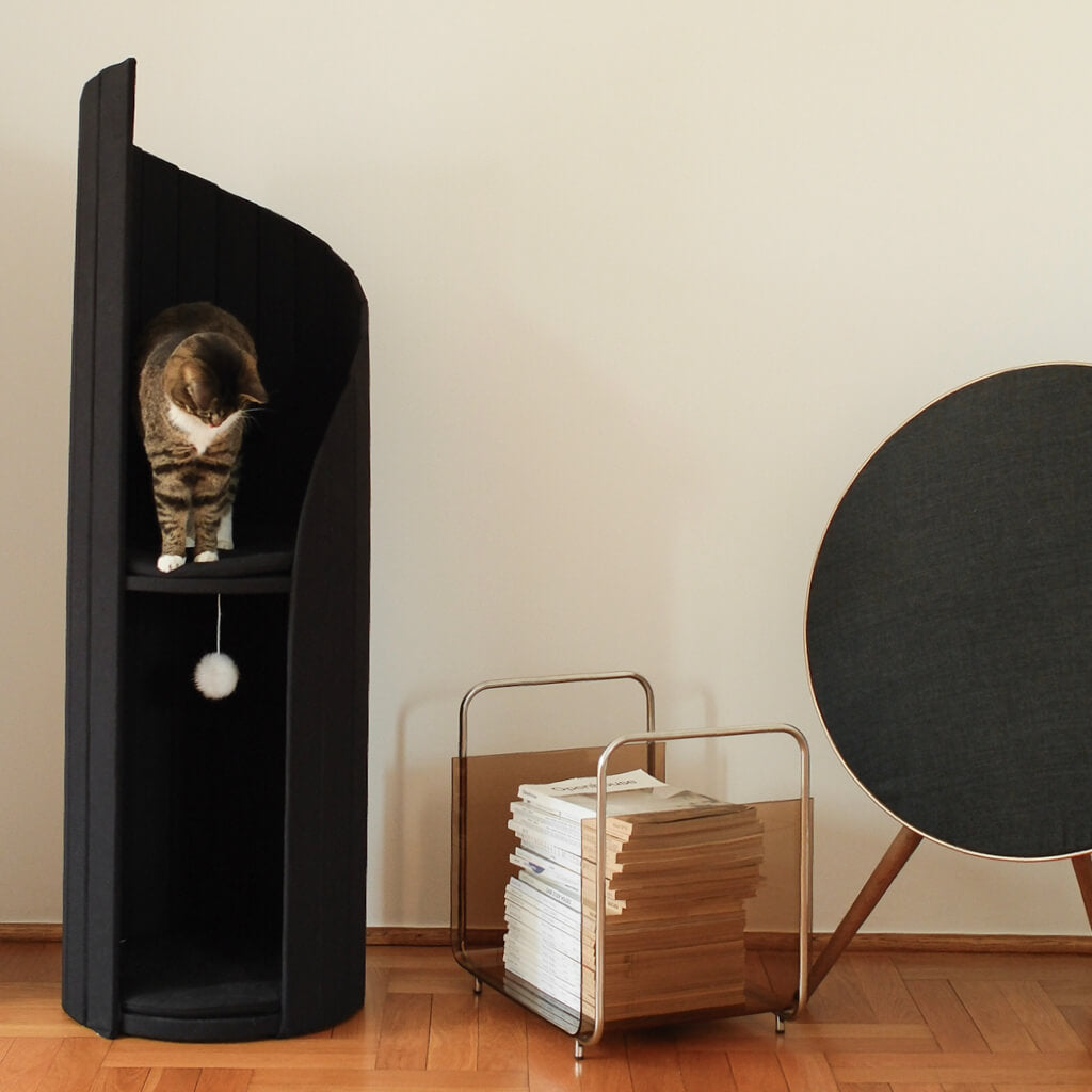 Tabby cat exploring the top level of the black Meoof Time Travel Cat Tower, next to a mid-century style magazine rack and speaker in a minimalist home setting.