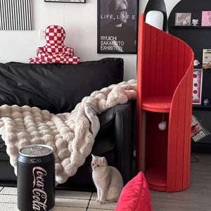 A modern living room setup featuring a black leather sofa, a cozy textured throw blanket, and a red Time Travel Cat Tower standing beside it. A curious light-grey cat sits on the floor, next to the tower, looking upward. The room is stylishly decorated with a Coca-Cola can-inspired furniture piece and framed artwork.