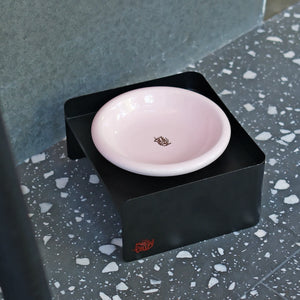 Black elevated pet bowl stand with pink ceramic dish – stylish metal base for cats and small dogs on speckled flooring.