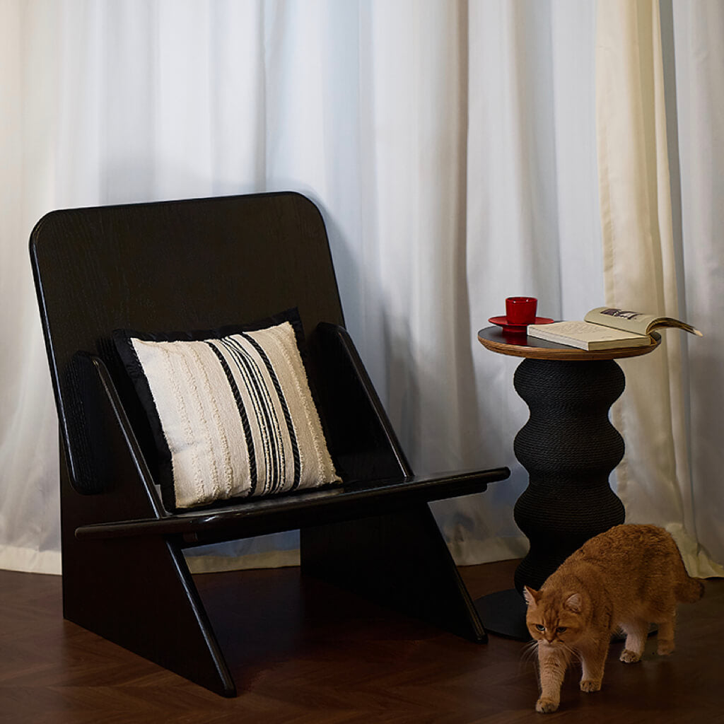Modern black wavy cat scratcher side table next to black lounge chair with decorative pillow, ginger cat walking by.