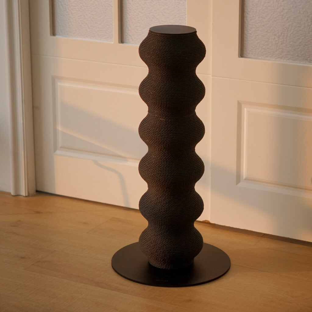 Black wavy cat scratching post in minimalist modern room, standing on light wooden floor with soft natural light.