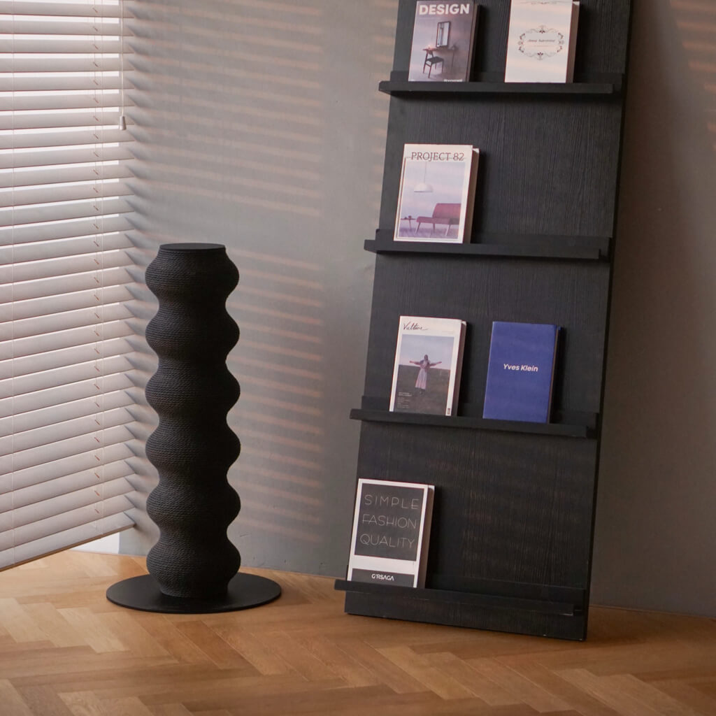 Modern black wavy cat scratcher next to magazine shelf and window blinds, styled in contemporary home corner.