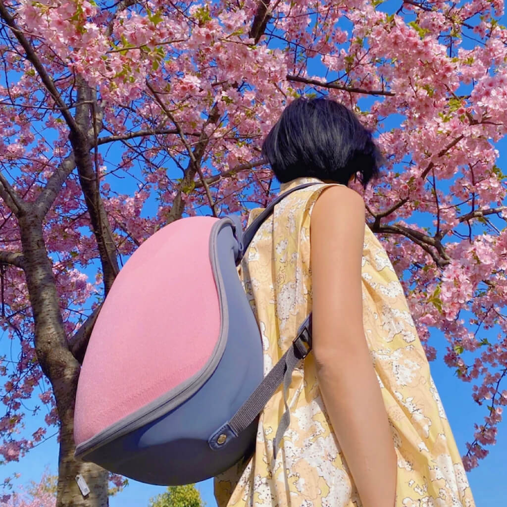 A person walking outside with the Extra-light Cat Travel Backpack - Pink+Grey, under blooming cherry blossoms, symbolizing travel and adventure.