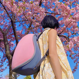 A person walking outside with the Extra-light Cat Travel Backpack - Pink+Grey, under blooming cherry blossoms, symbolizing travel and adventure.