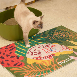 Cat stepping out of green litter box onto Purrre Tropical Jungle Cat Mat – colorful litter mat with jungle leaves and lounging cat illustration.