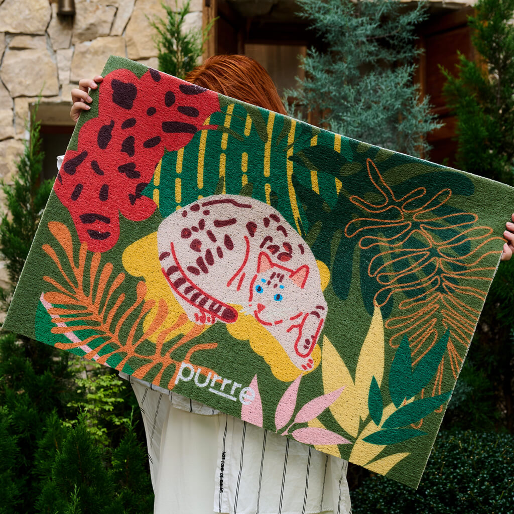 Woman holding up large colorful cat litter mat – jungle foliage print and sleeping cat graphic.