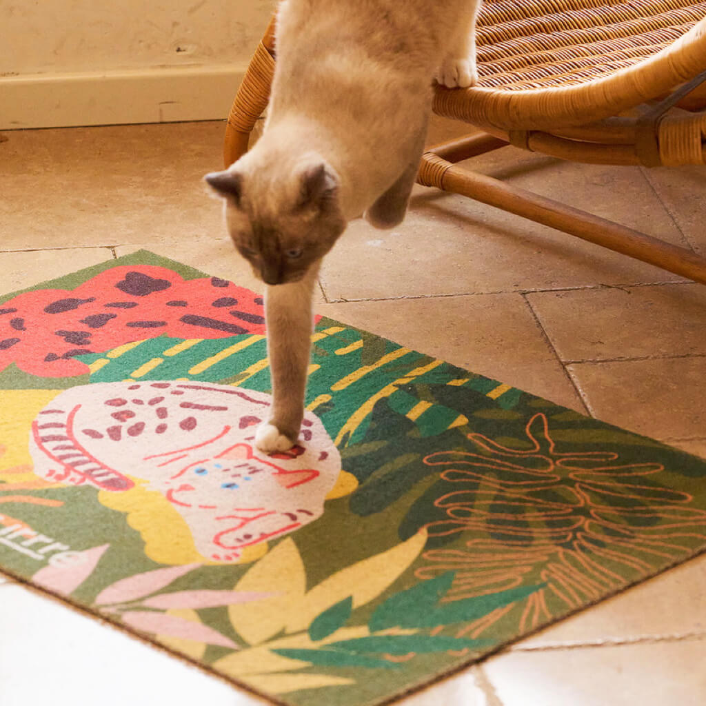 Cat walking off rattan chair onto soft Purrre cat mat with tropical greenery and feline print.