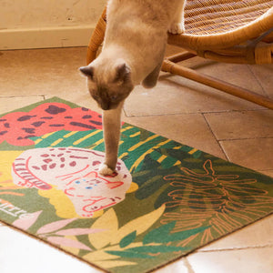 Cat walking off rattan chair onto soft Purrre cat mat with tropical greenery and feline print.