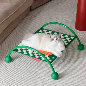 Fluffy white cat lounging on a vibrant green Memphis style elevated hammock with checkerboard pattern, set against a domestic living room background.