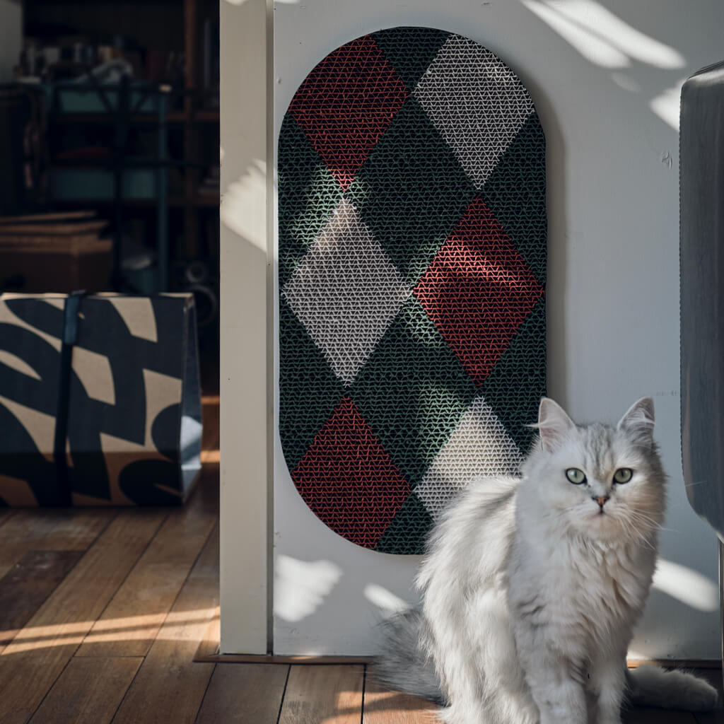 Cat sitting next to wall-mounted Checker cat scratcher with a red, green, and white pattern.