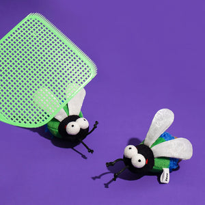 Catnip fly toy under a mint green swatter with another bug toy beside it on a purple background.