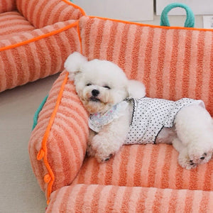 Small white dog resting in orange striped plush cat bed with soft plush fabric.