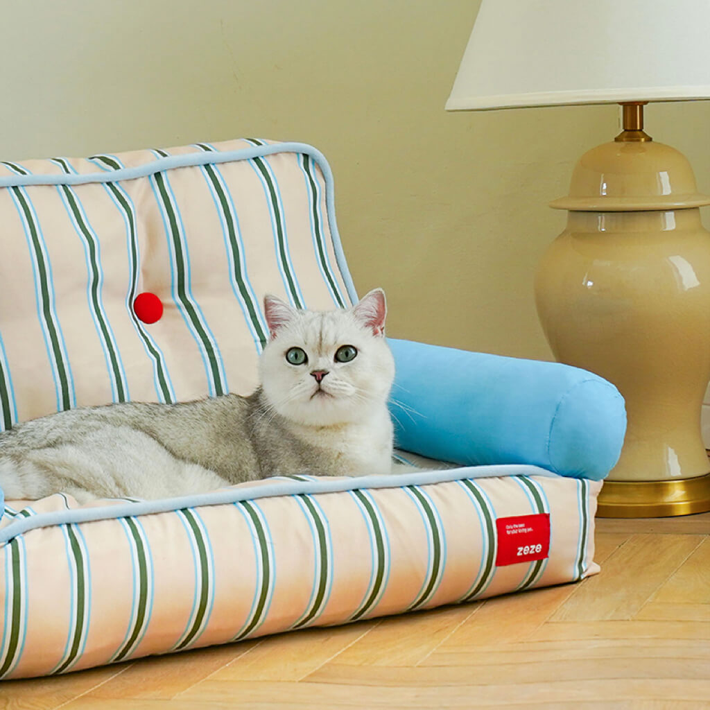 Cat sitting comfortably on ZEZE striped pet sofa next to a beige table lamp
