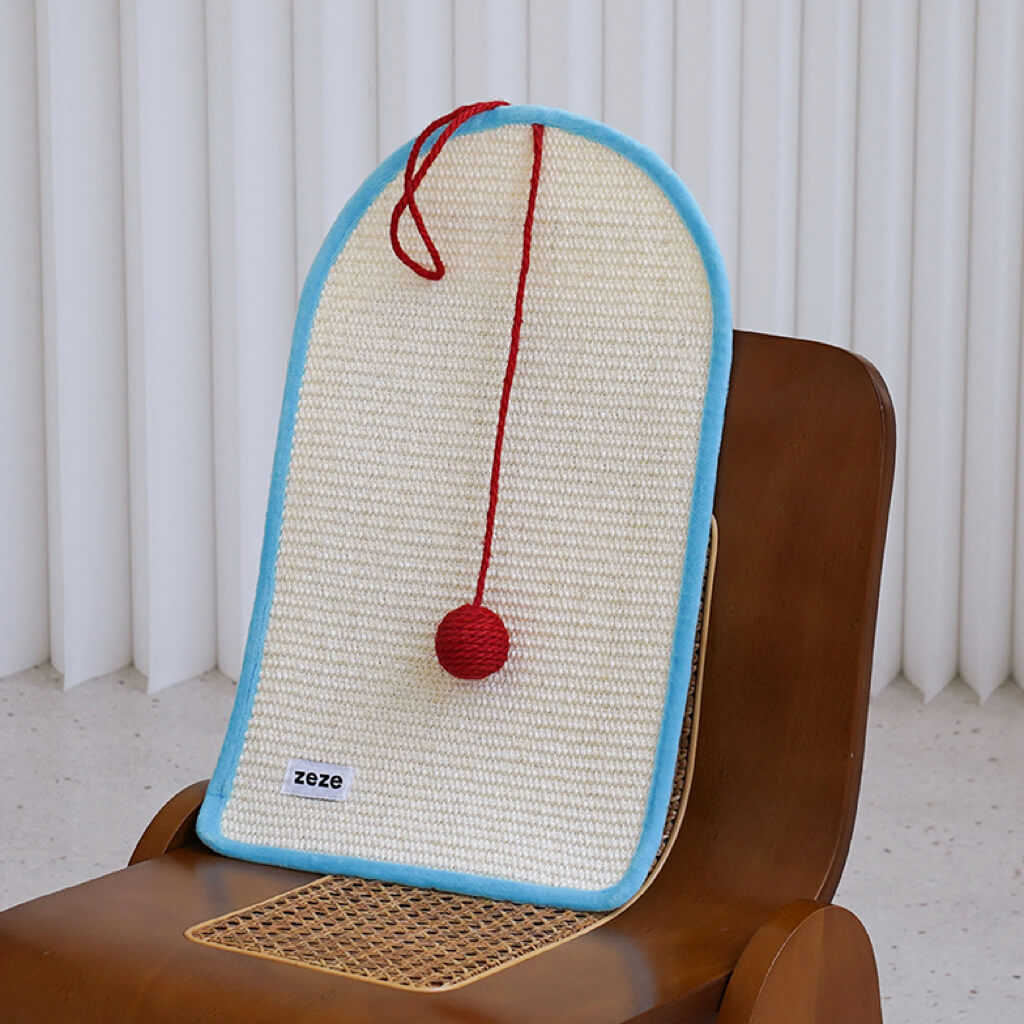 Modern cat scratching mat with red pom-pom and blue edge on a design chair.
