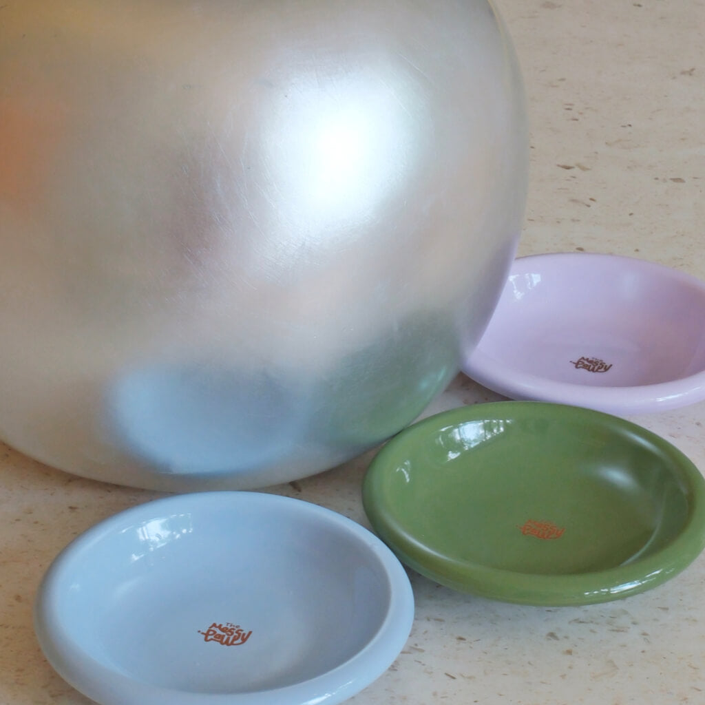 Stackable Messy Paws shallow ceramic cat bowls in pastel blue, green and purple styled beside a silver orb on a stone floor—modern home décor.