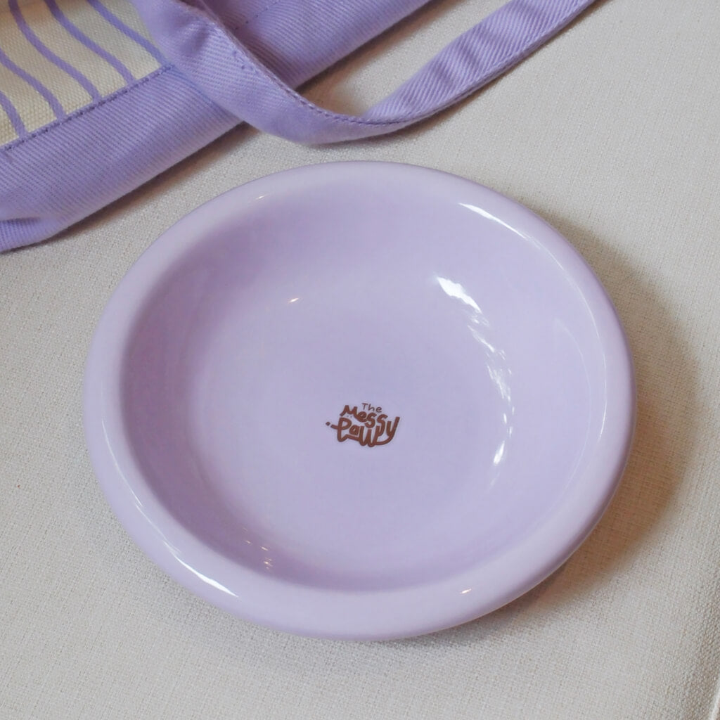 Light purple Messy Paws shallow ceramic cat bowl on fabric next to a lavender tote strap—glossy, minimalist pastel pet dish by Pewos.