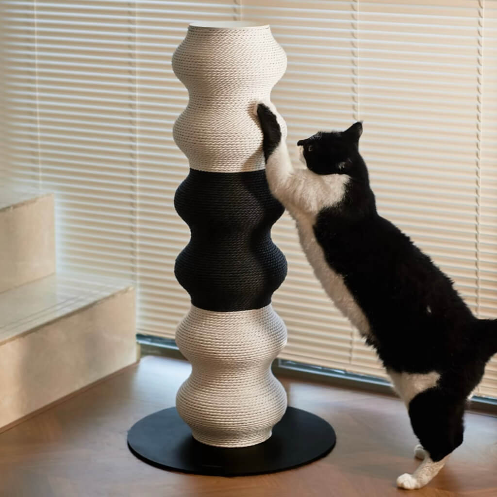 Black-and-white cat stretching on the Coral Cat Scratcher by window blinds; wavy post with wide steel base.