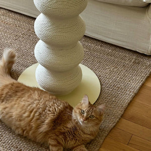 Close-up of White Momoland Coral Scratcher – wavy vertical cat scratching post with round base; ginger cat relaxing on rug next to durable floor-standing scratcher.