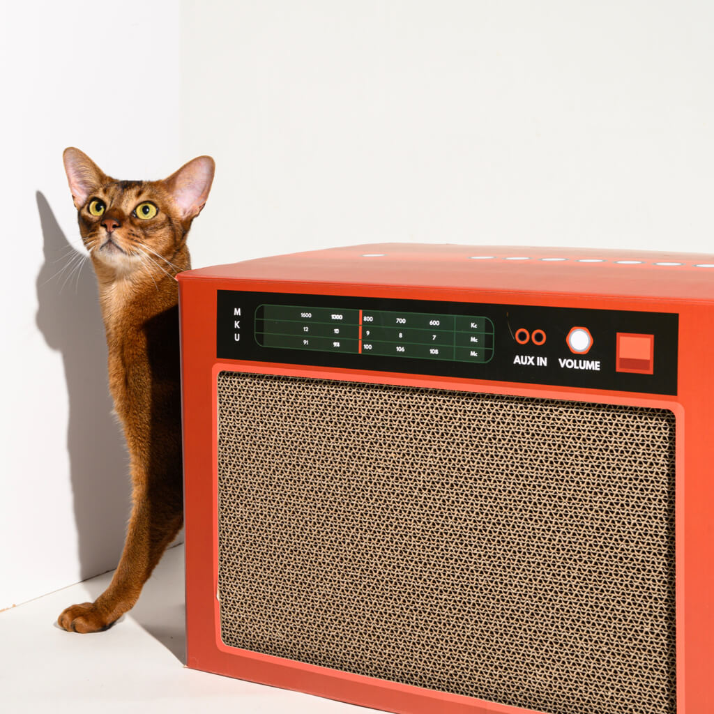 Brown cat standing beside red Pawzcity stereo scratcher – multi-functional design with durable corrugated cardboard.
