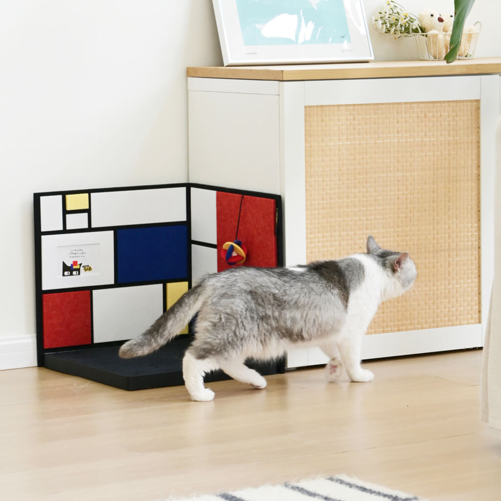 Corner cat scratcher in modern living room — L-shaped, color-block panels with base; cat walking nearby; Mondrian-inspired design.