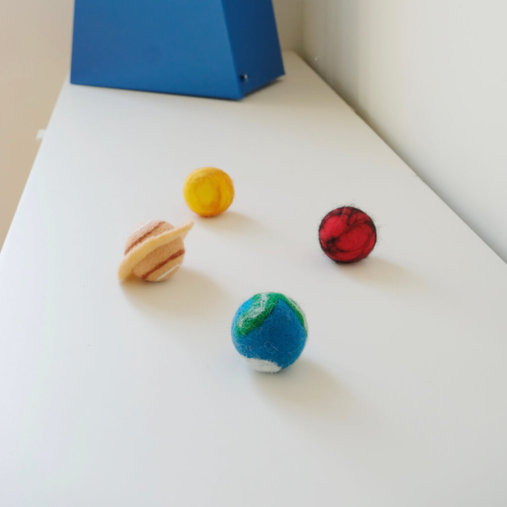 Four solar-system felt cat balls on a white shelf — Earth, Mars, Sun, Saturn.