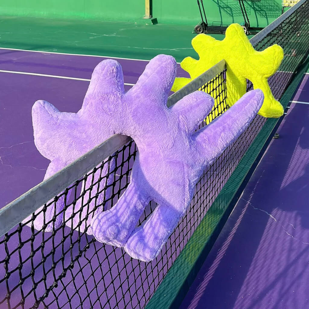 Gelato Purple and Fluo Green Boom Boom Cat Cushions perched on a tennis net.