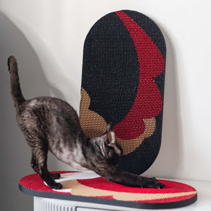 Cat is scratching on Hand-Crafted Paper Art Cat Scratcher in red, white, beige, black. One is laying on the ground, other is mounted on a wall.