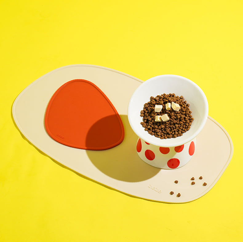 Beige and orange silicone placemat with a pet food bowl filled with kibble on a bright yellow background.