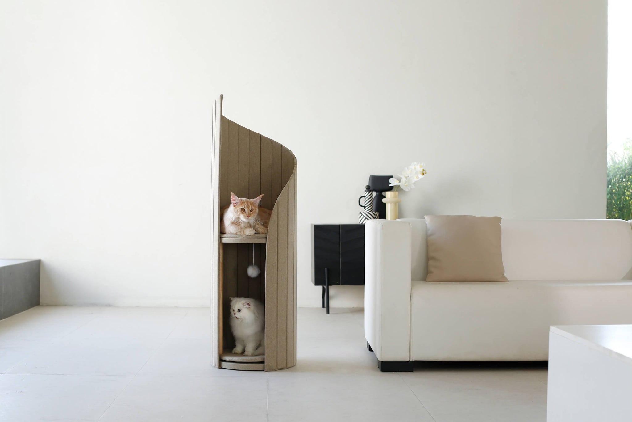 Ginger and white cats relaxing on different levels of the beige Time Travel Cat Tower, placed next to a modern white sofa in a minimalist living room.