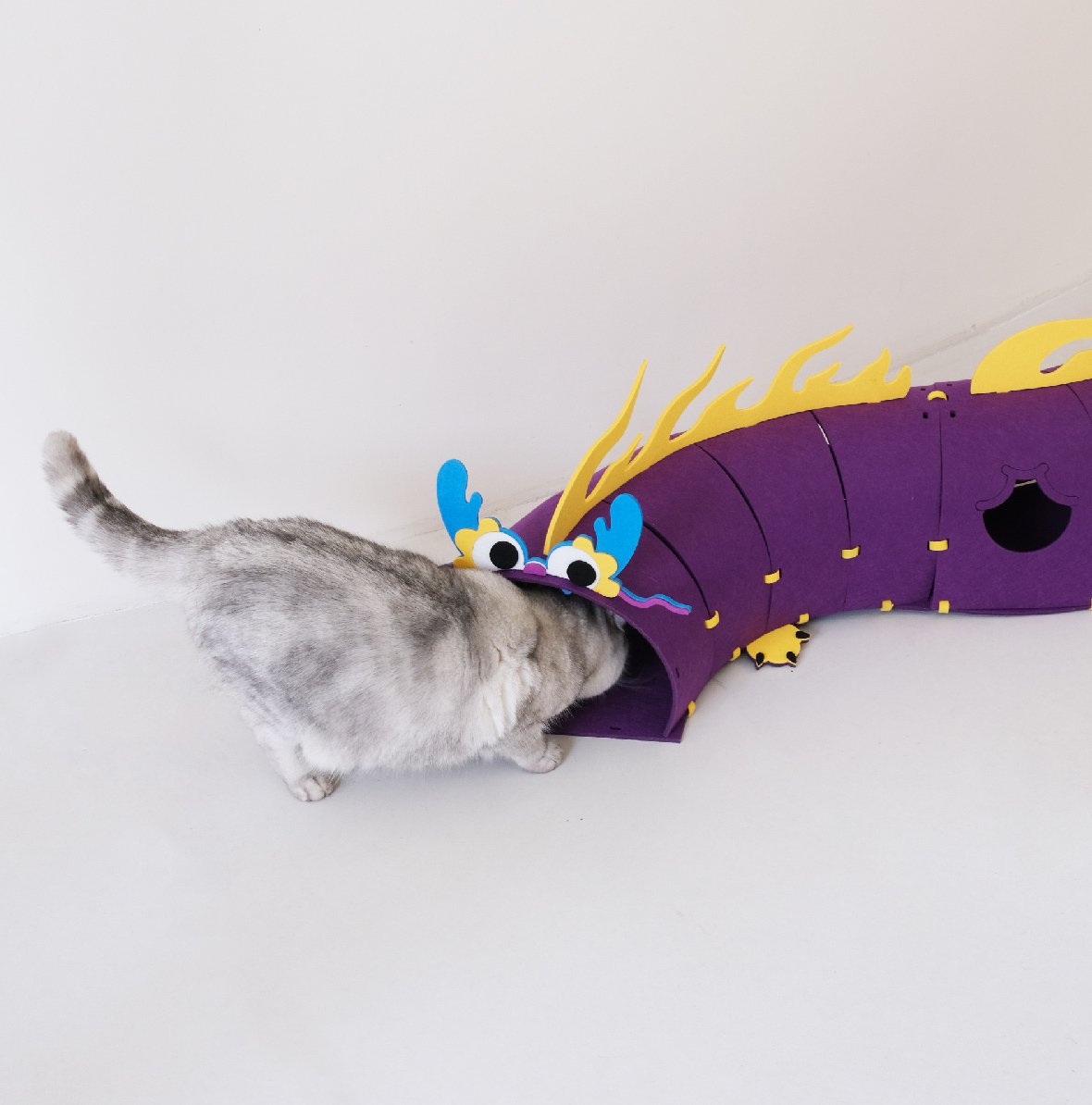 Cat entering a purple felt dragon tunnel with yellow flames and blue accents - Design Cat Felt Tunnel Toy - Dragon Edition.